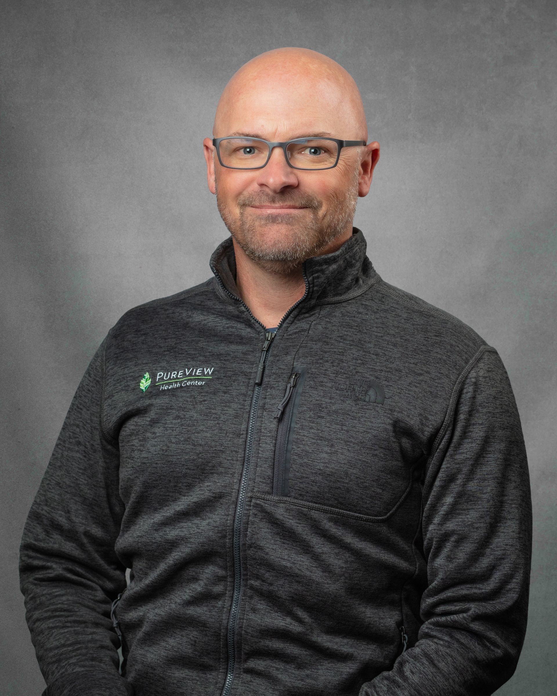 A bald man with glasses and a beard is smiling in front of a brick wall.