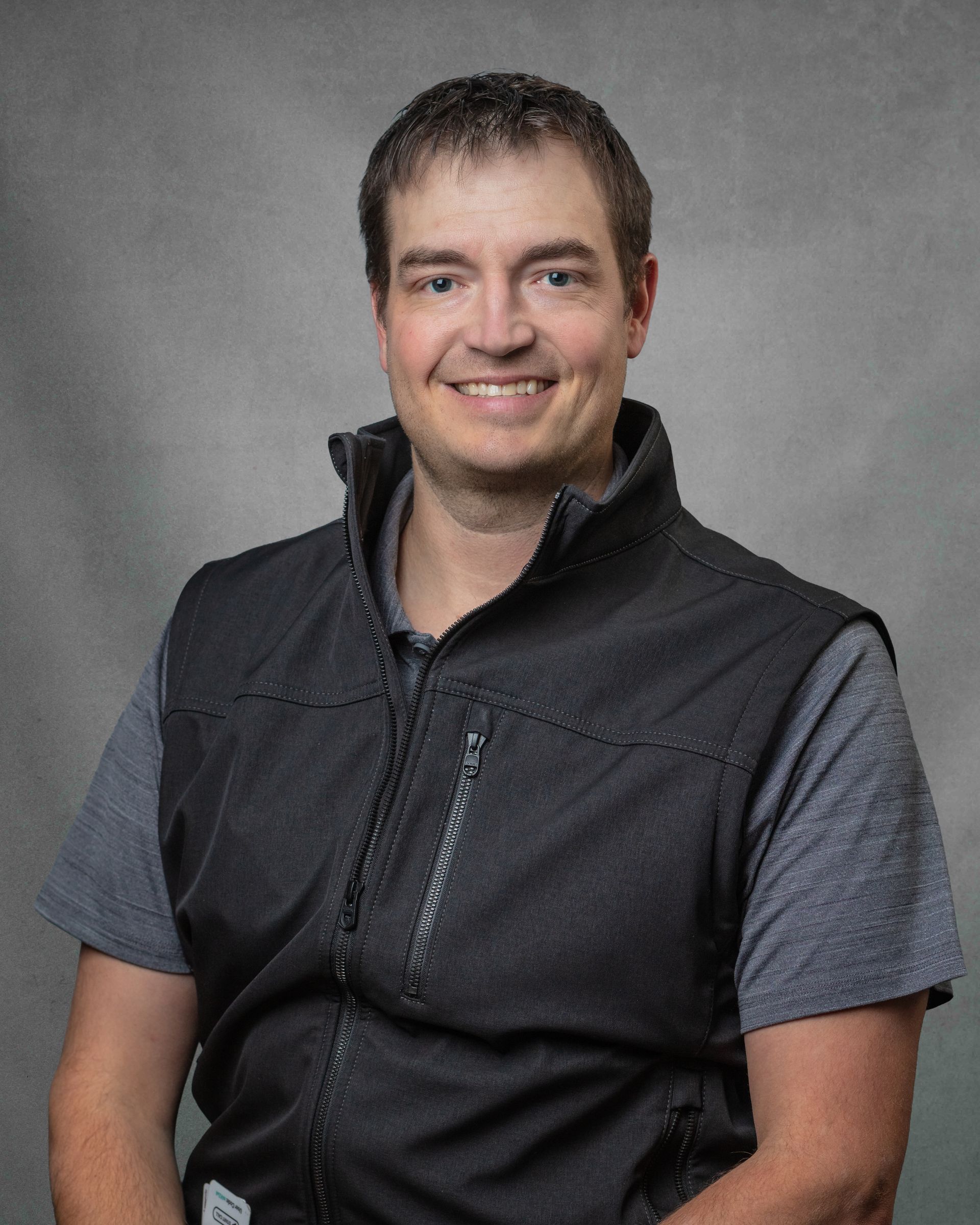 A man in a black vest is smiling in front of a gray background