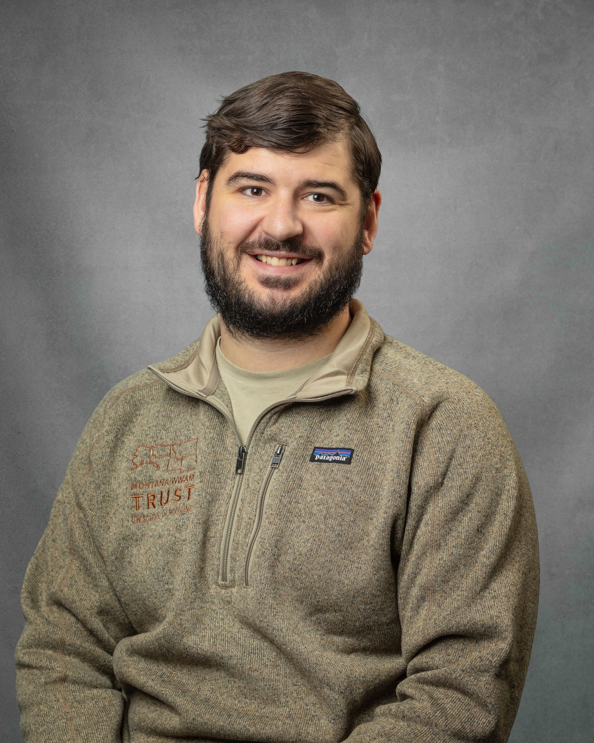 A man with a beard in a brown zip up is smiling.