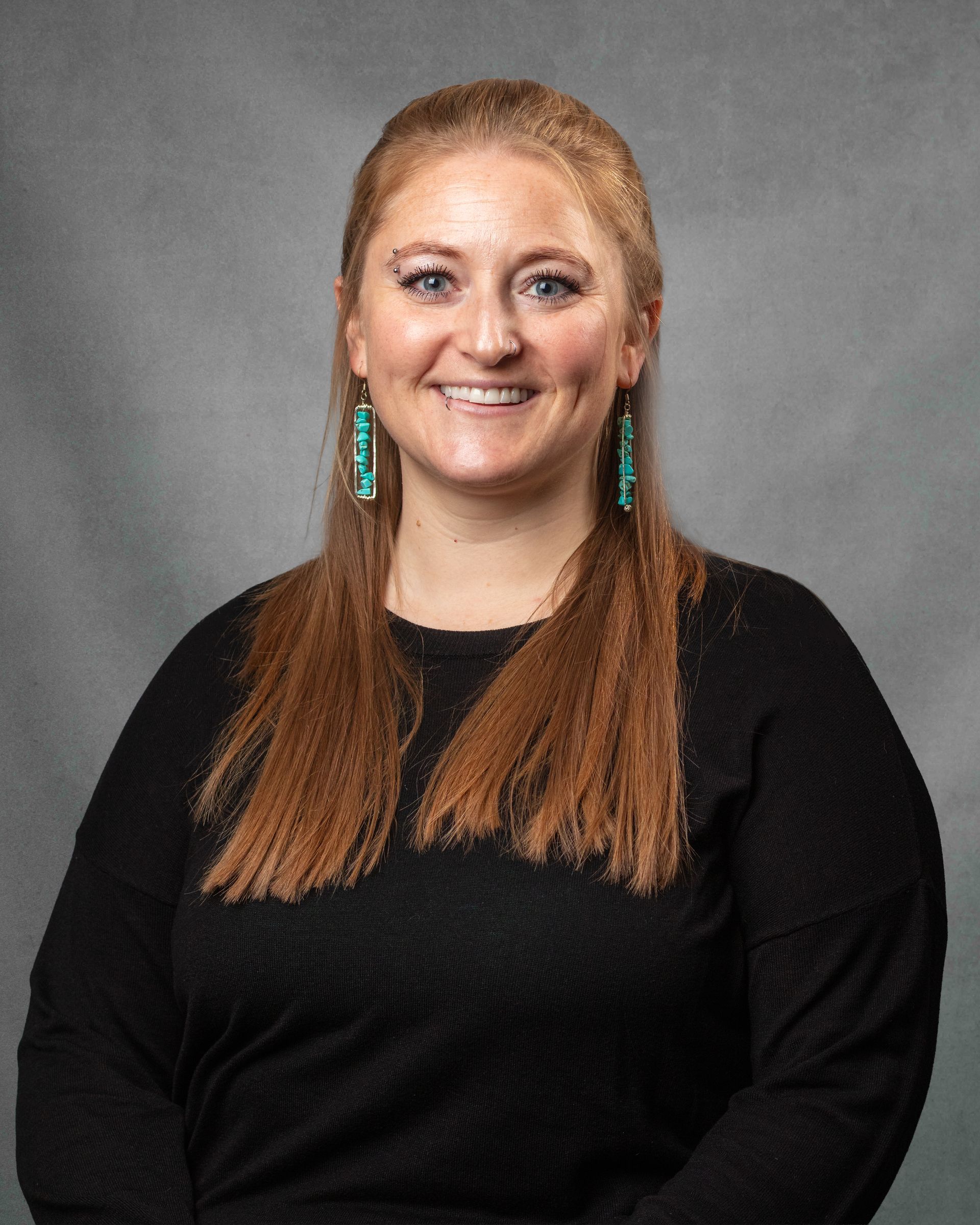 A woman wearing black shirt with blue earrings and an eyebrow piercing is smiling for the camera.