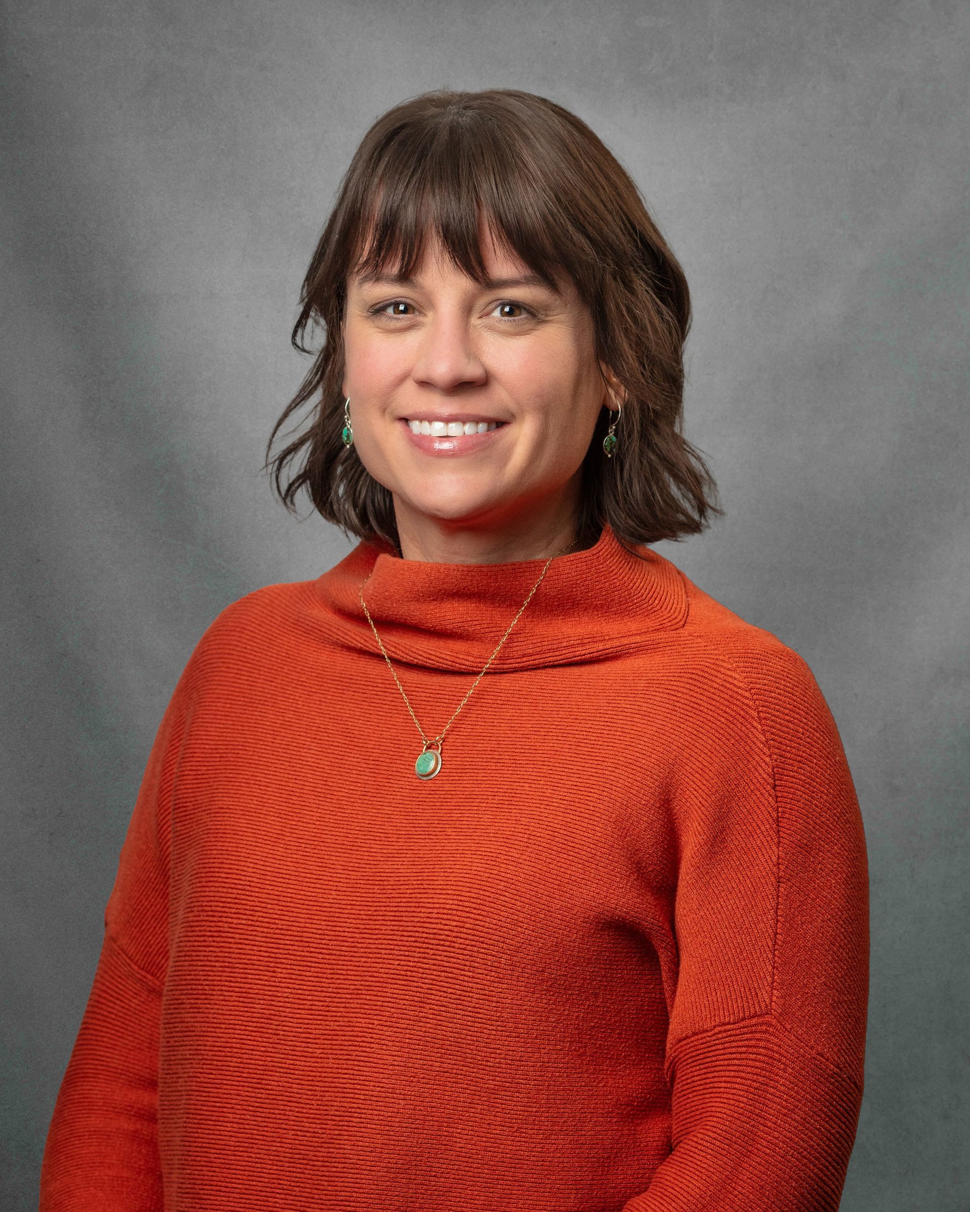 A woman in a green jacket is smiling in front of a brick building.