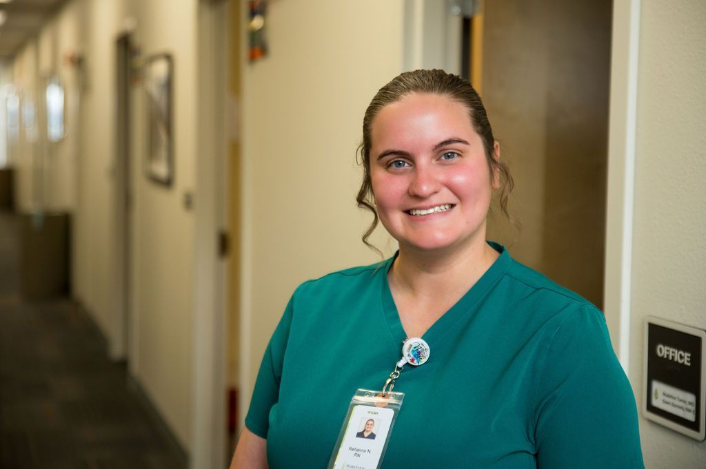 A woman in a green scrub is standing in a hallway and smiling.