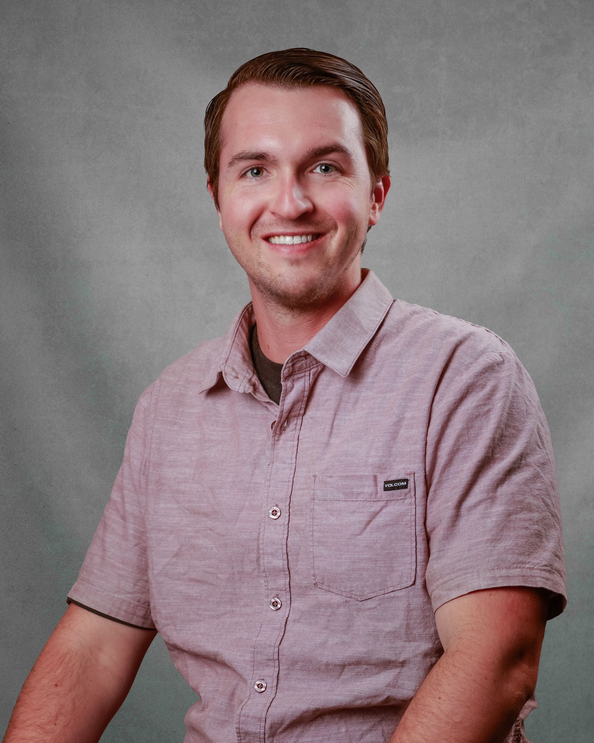 Smiling man in a pink short-sleeve shirt seated against a gray studio backdrop