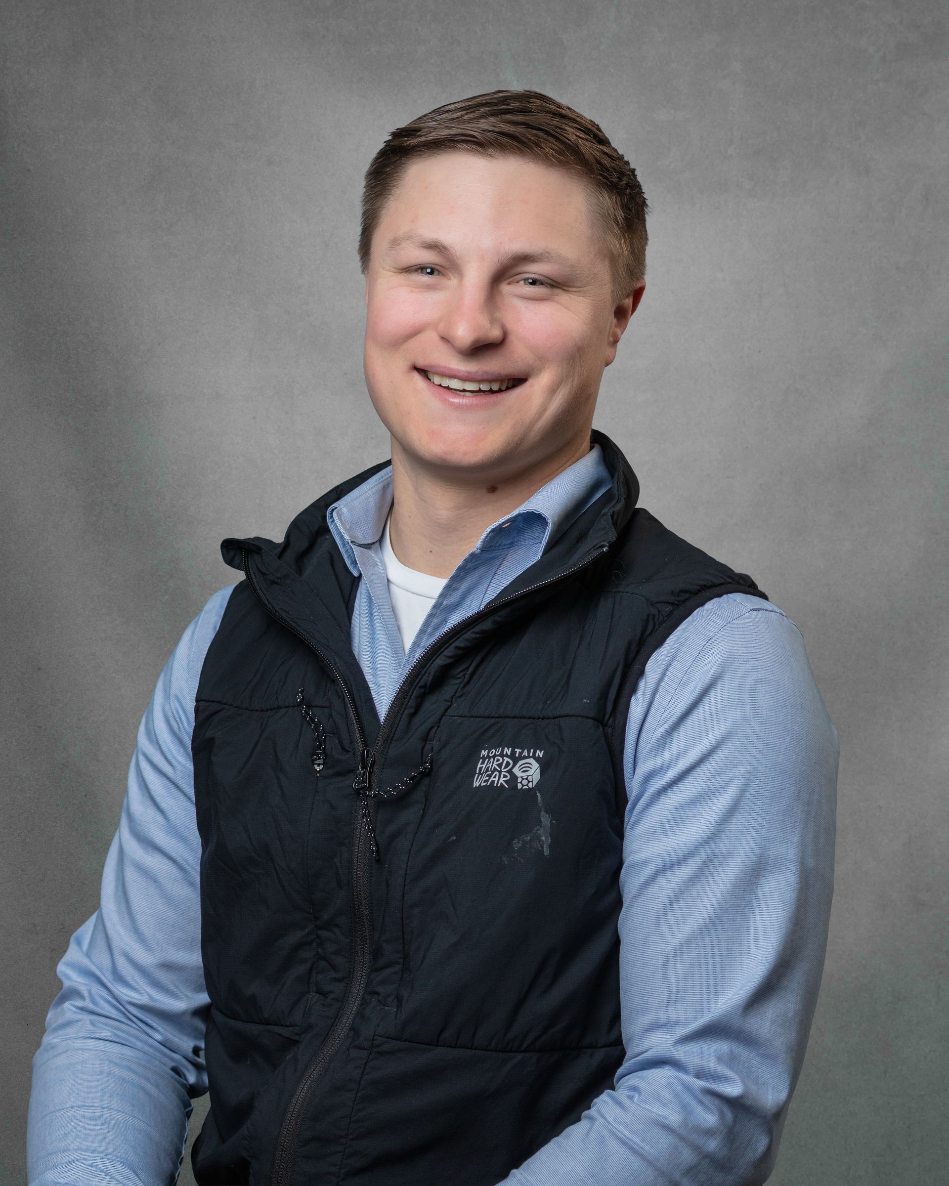 A man in a blue shirt and a black vest is smiling.