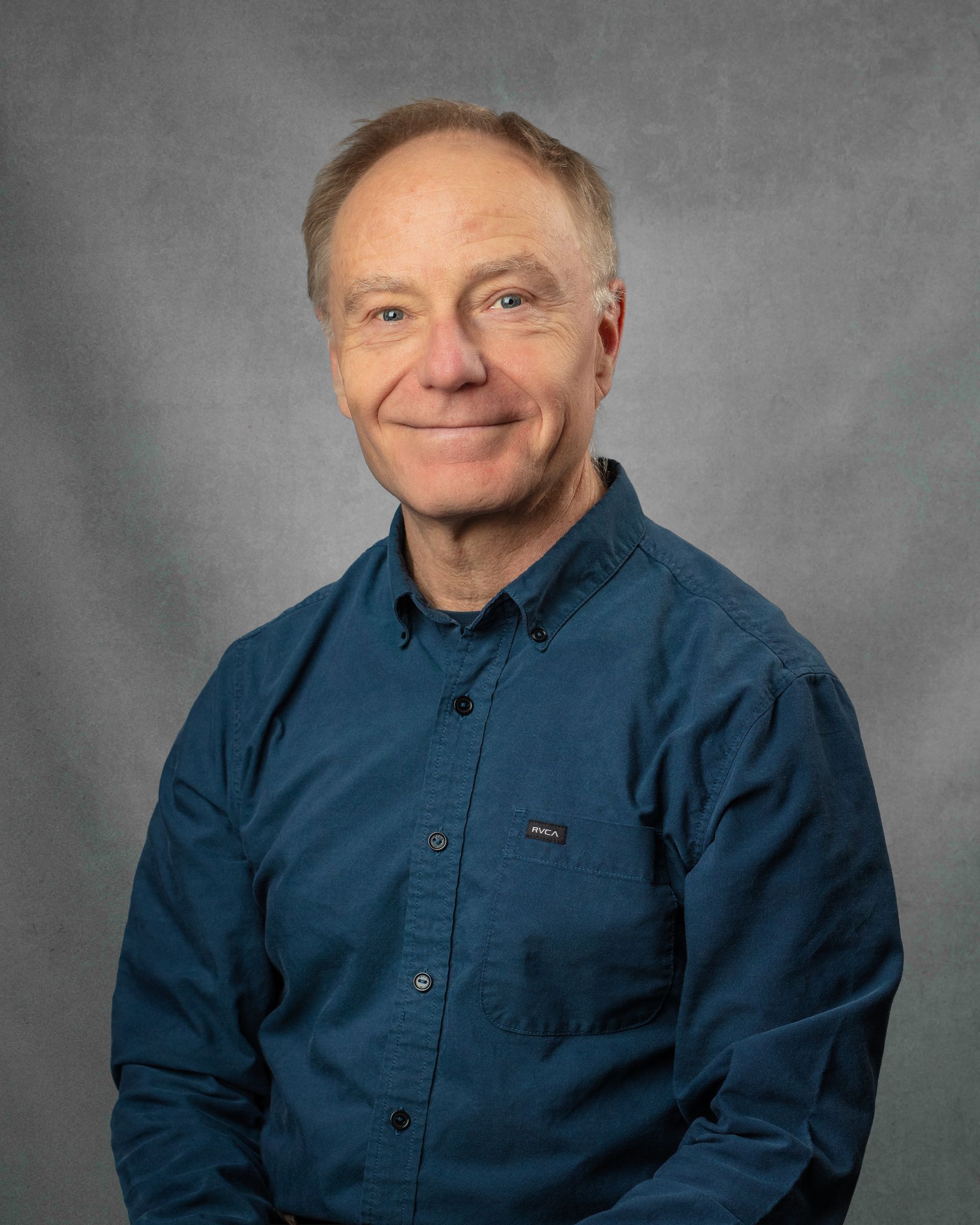 A man wearing a blue button up shirt is smiling in front of a brick wall.