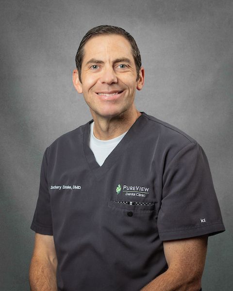 A man in a scrub top is smiling in front of a gray background.