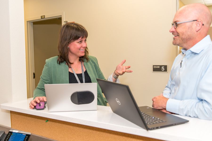 A woman is talking to a colleague at a desk in front of a computer.