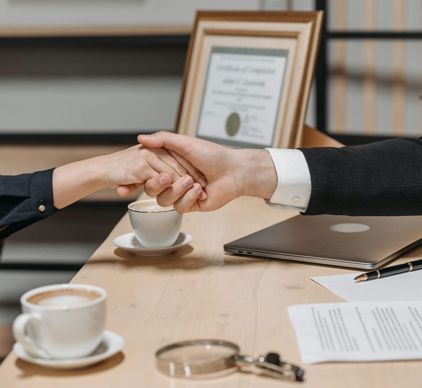 Two people shaking hands across a desk with coffee cups and a laptop in a meeting room