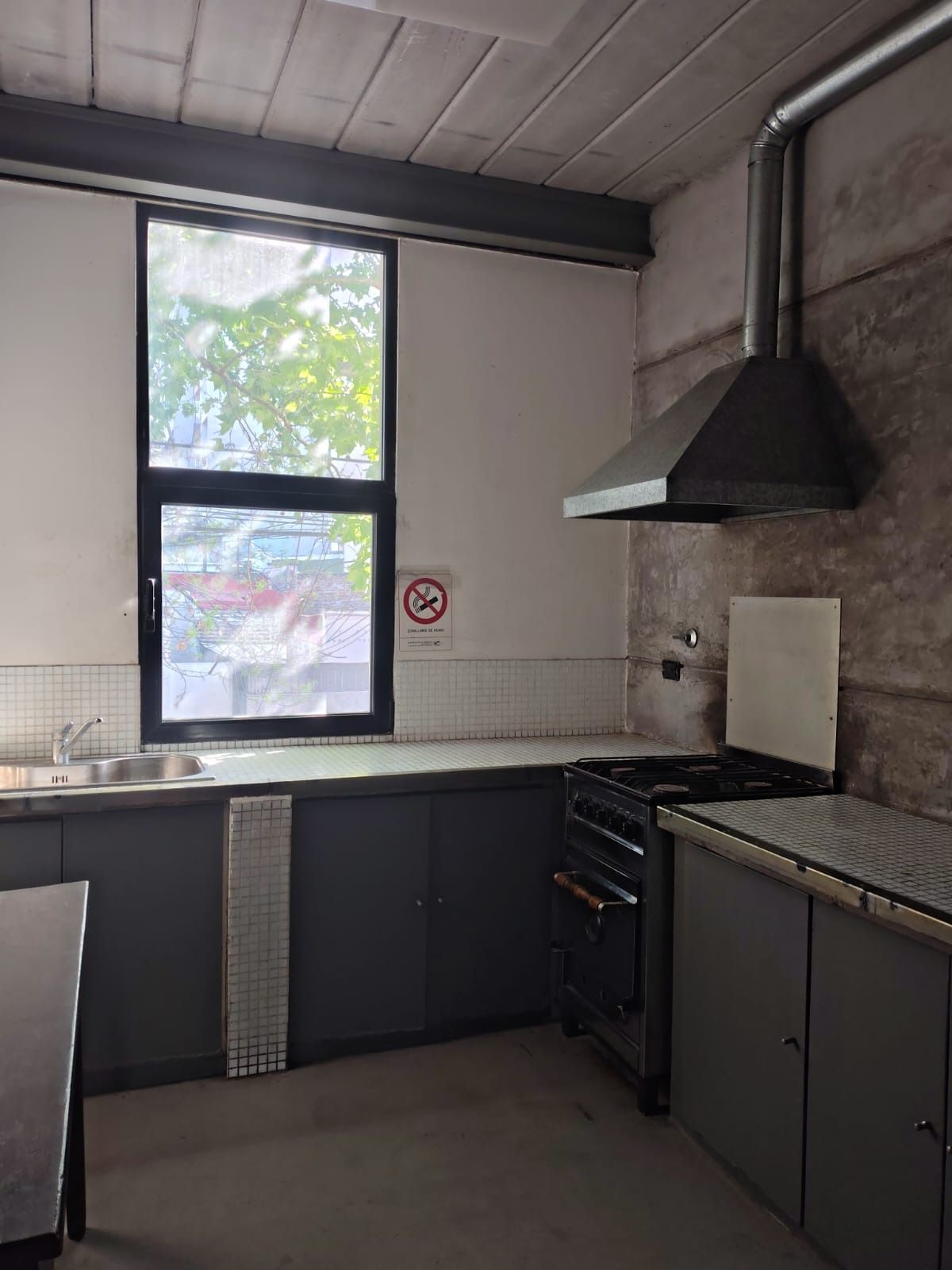 Cocina pequeña con armarios oscuros, paredes de azulejos blancos, ventana, fregadero, estufa y campana extractora negra.