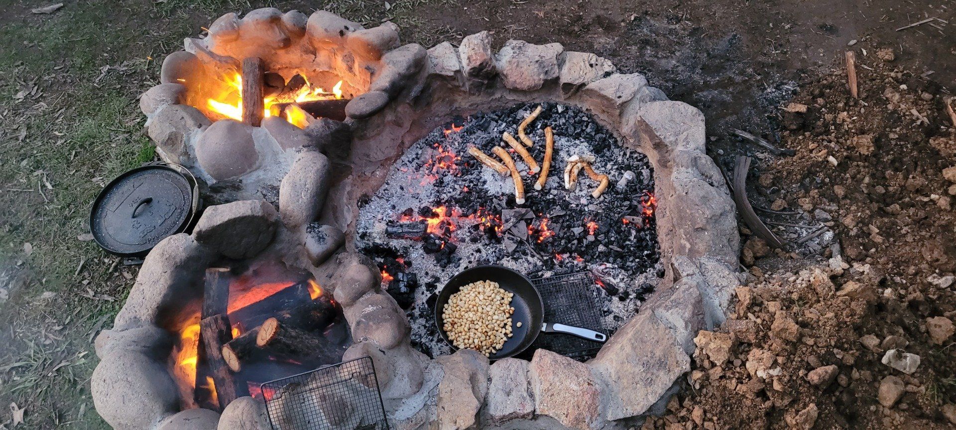 A stone fire pit with burning wood and cooking food, including a pan of pasta.