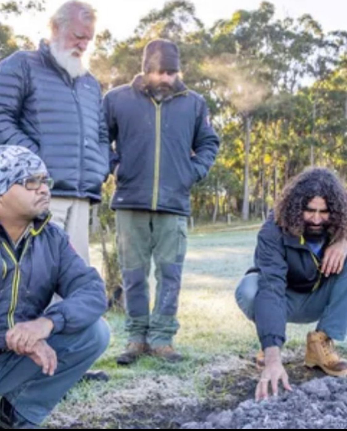 Four people outdoors, examining soil. One squats, touching the ground. Cool air visible.
