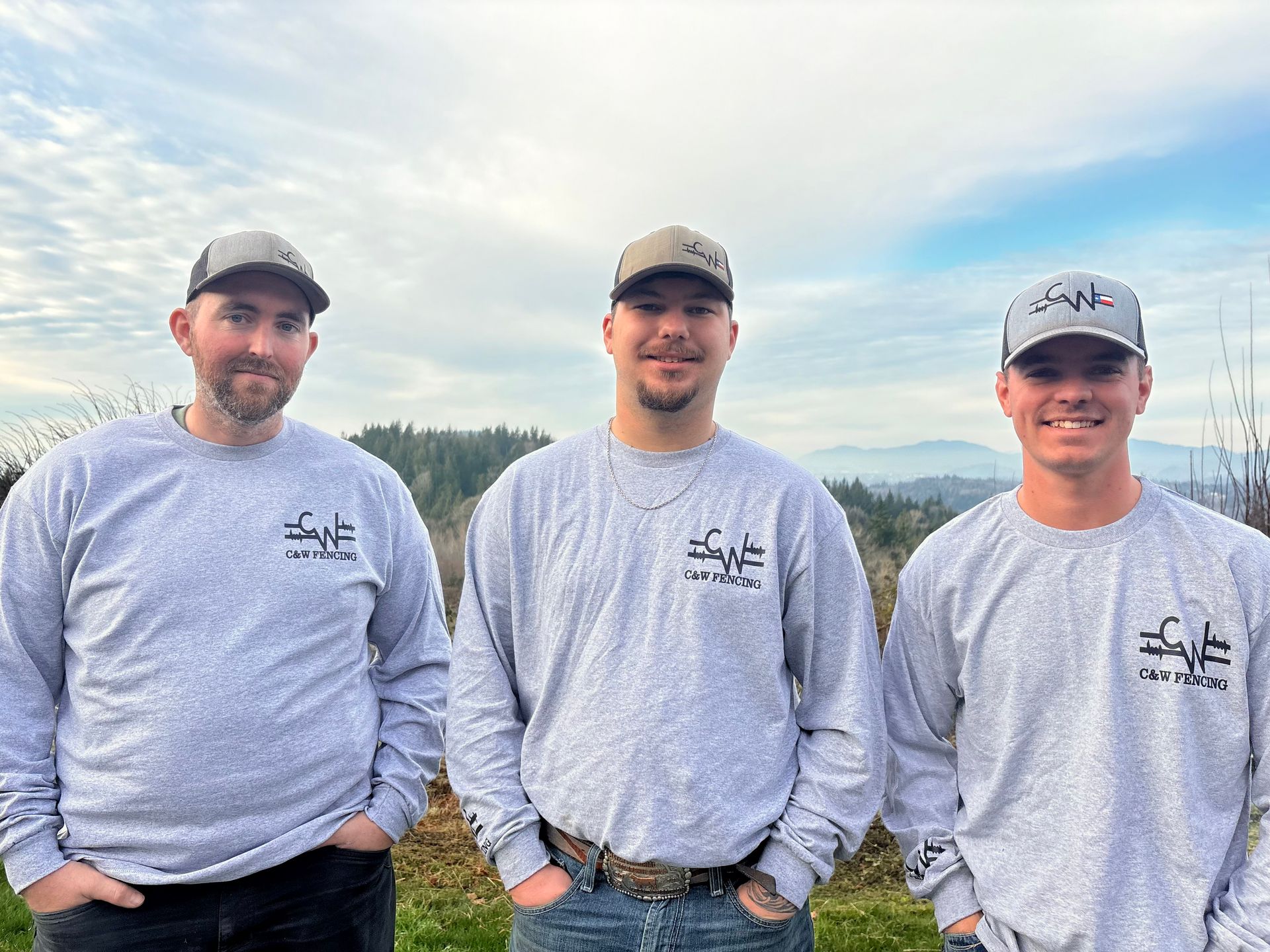 Three people in gray shirts standing outdoors under a cloudy sky, smiling at the camera.