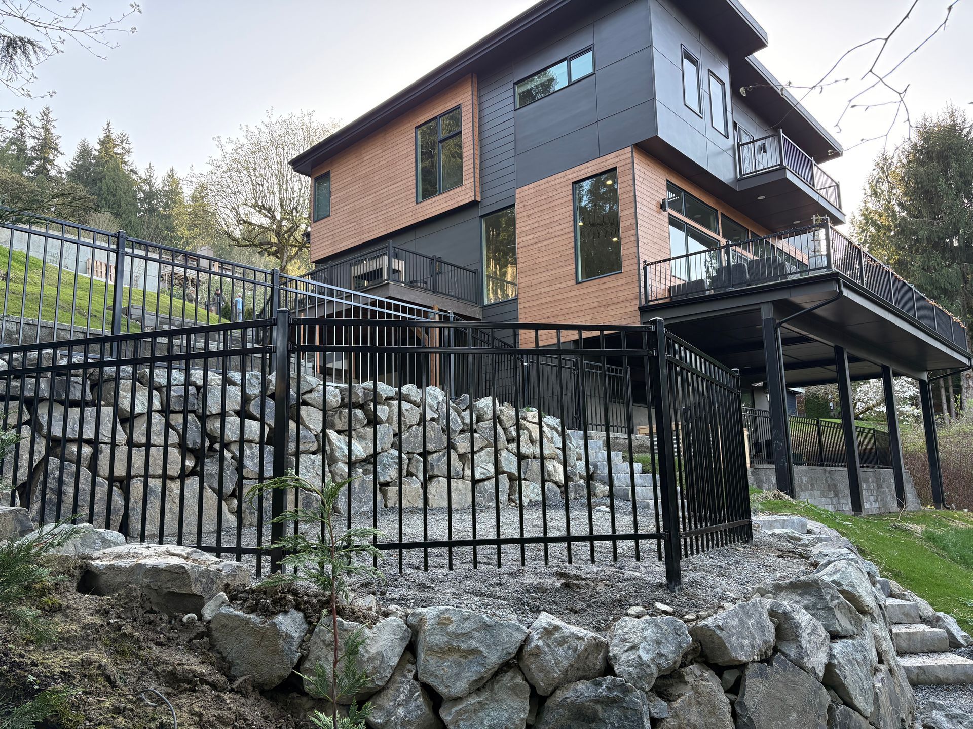 Modern house on a steep stone slope behind a black metal fence, with trees in the background.