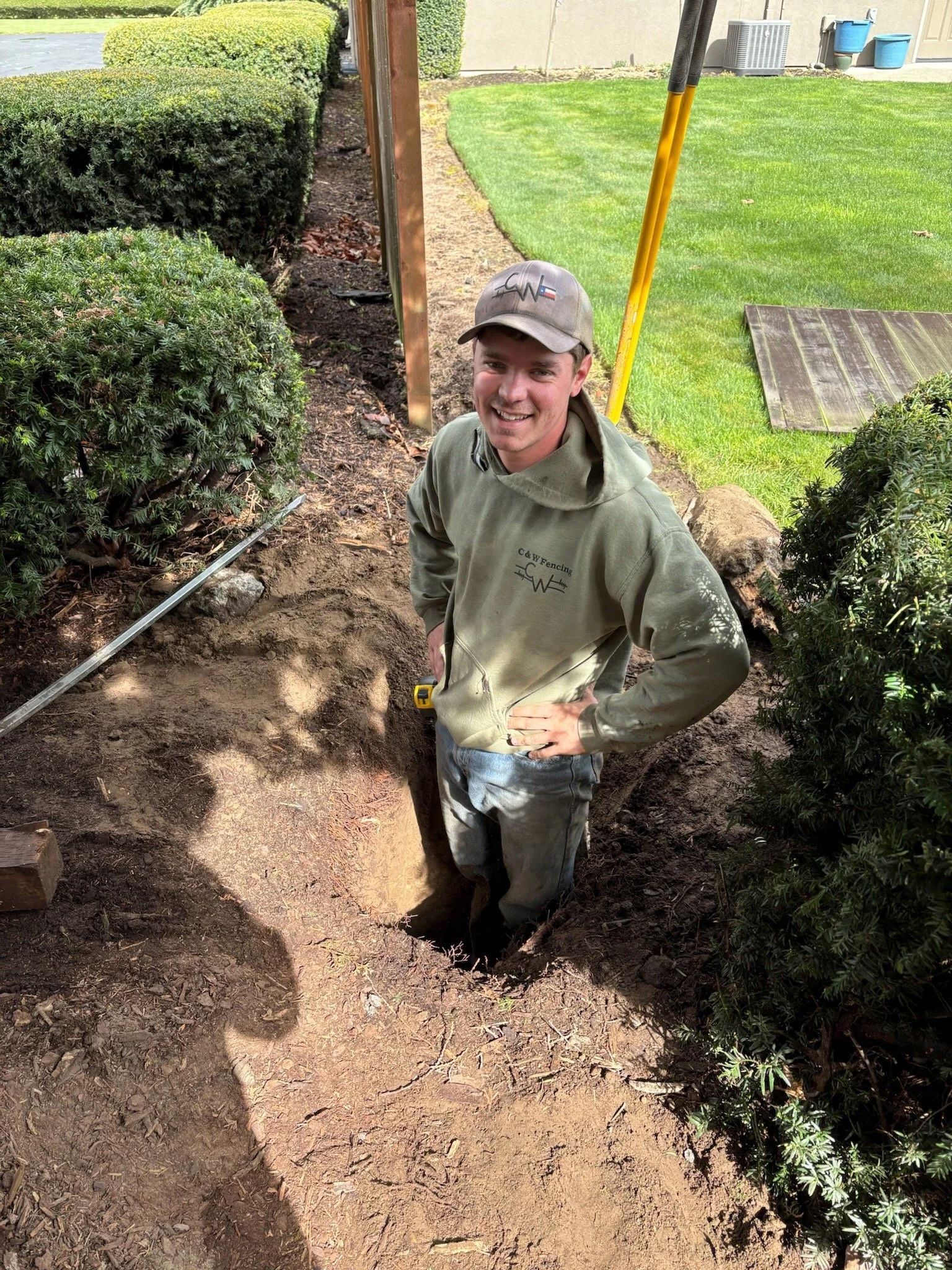 Person standing in a dirt trench beside hedges, smiling and holding a shovel in a yard.