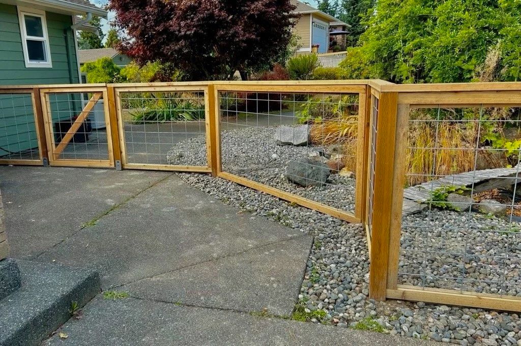 Wooden fence with wire panels lining a gravel yard beside a paved driveway and house garden