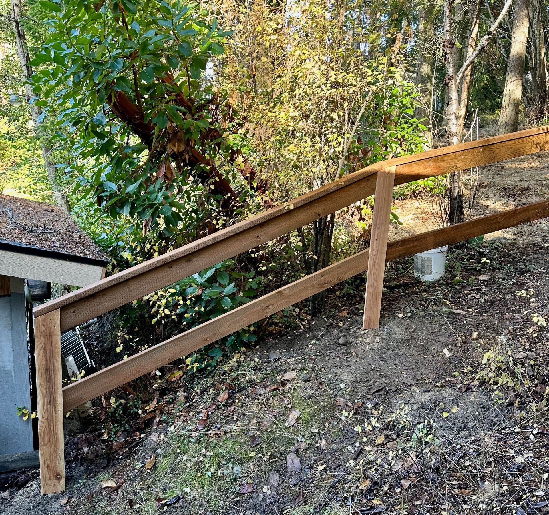 Wooden handrail along a sloped dirt path in a wooded yard.