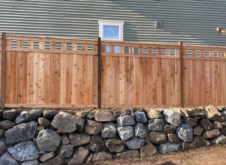 Wooden privacy fence atop a stacked stone retaining wall in front of a house roofline