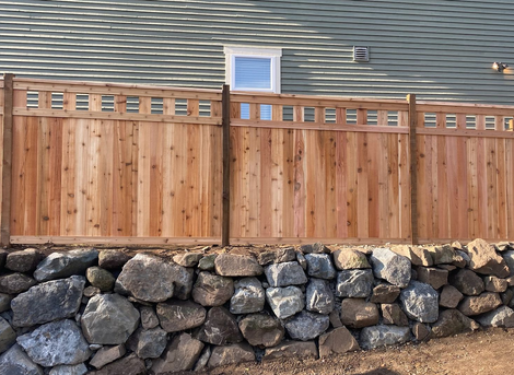 Wooden privacy fence atop a stacked stone retaining wall in front of a house roofline