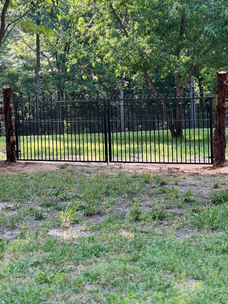Black metal gate between stone posts in a leafy yard, with a grassy foreground and trees beyond.