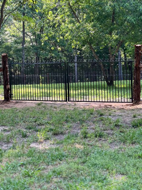 Black metal gate between stone posts in a leafy yard, with a grassy foreground and trees beyond.