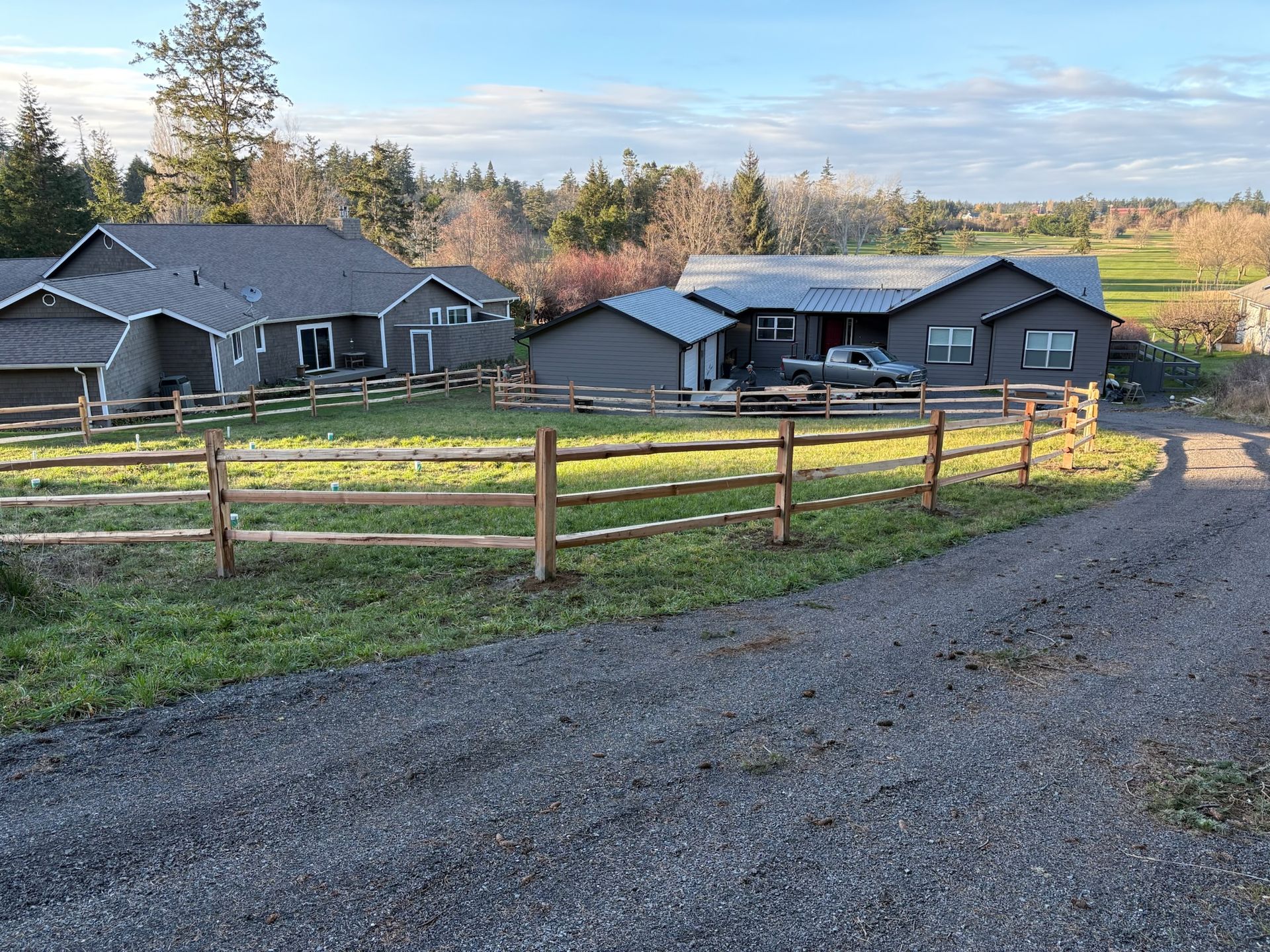 Rustic homes behind a wooden fence beside a gravel driveway, with trees and open fields under a clear sky.