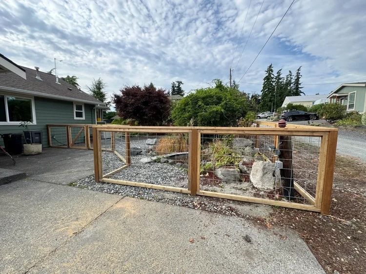 Wooden framed garden enclosure beside a driveway with rocks, plants, and a stone birdbath