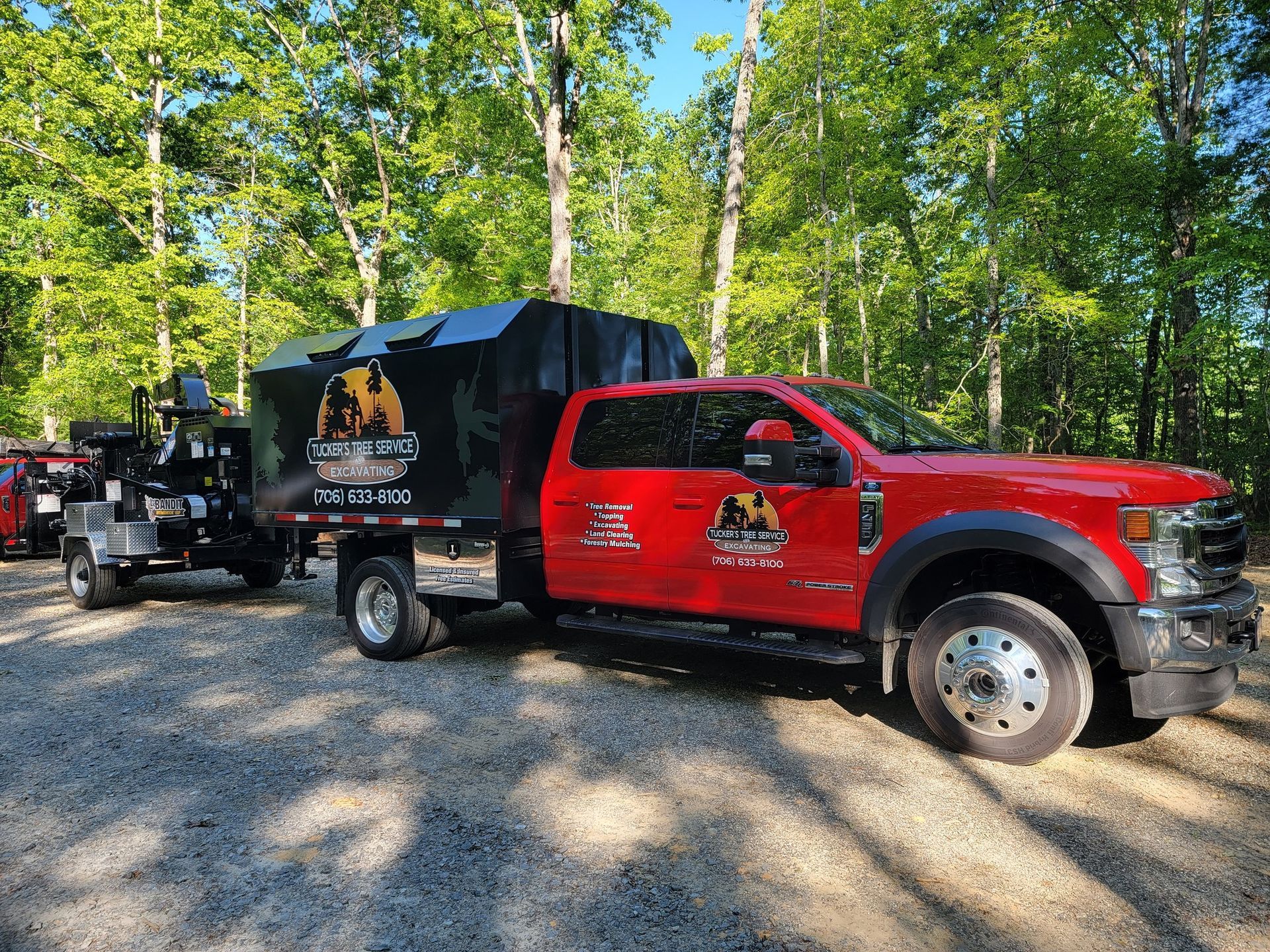 A red truck is parked in a gravel lot with trees in the background.