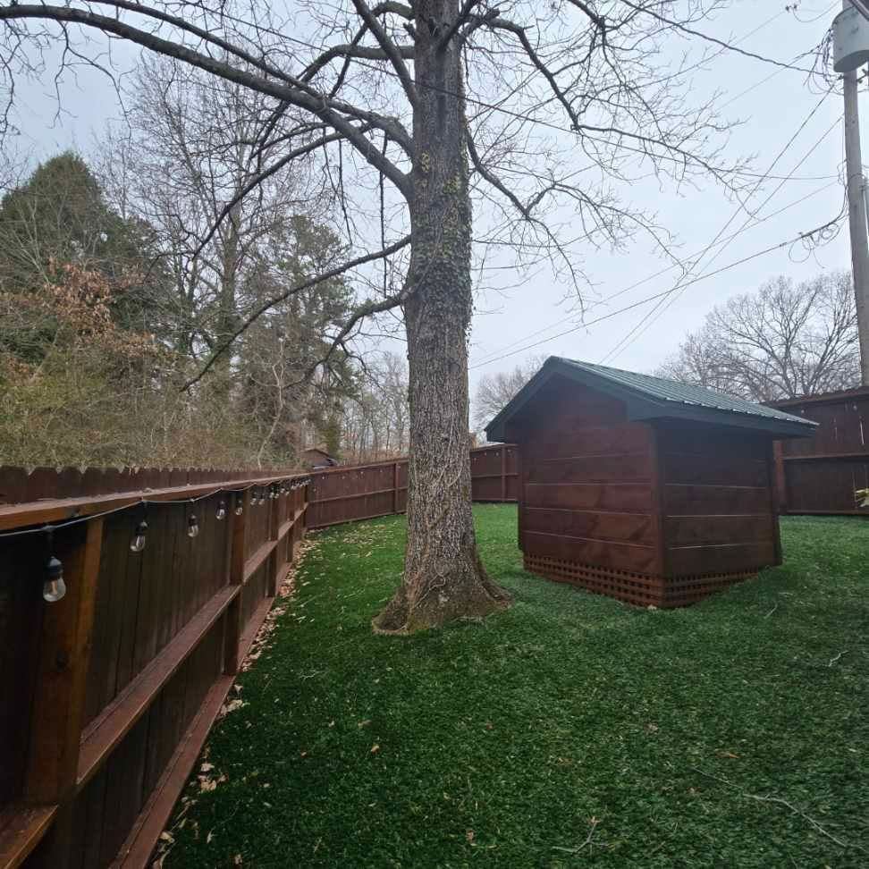 A wooden shed is sitting in the middle of a lush green yard next to a wooden fence.