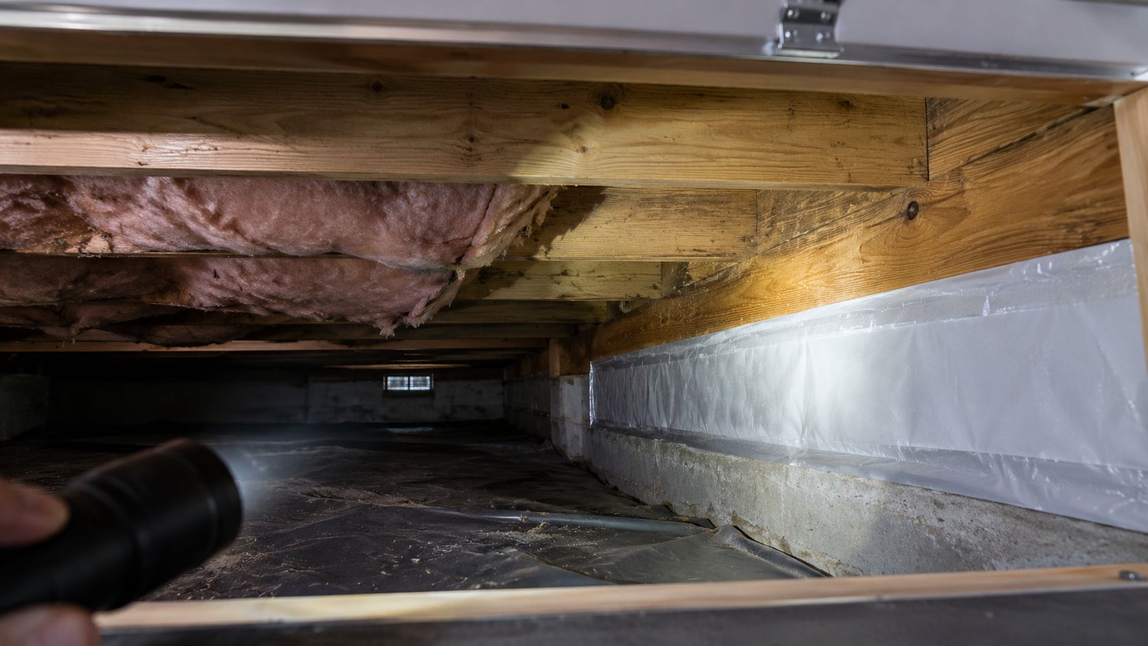 Crawlspace with exposed insulation, wooden joists, and plastic vapor barrier along the wall