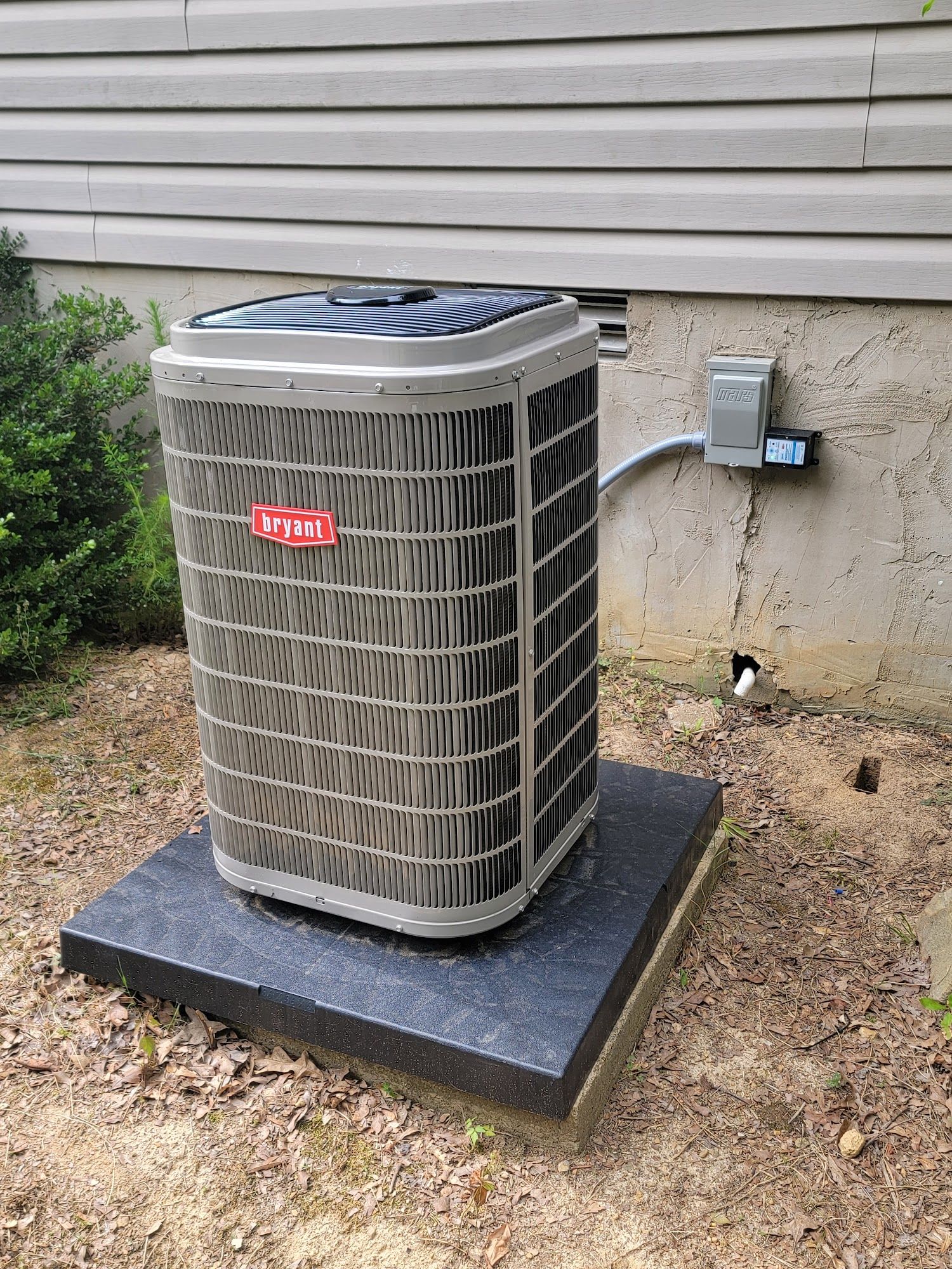 Outdoor air conditioner unit on a concrete pad beside a house wall, with a power box nearby.