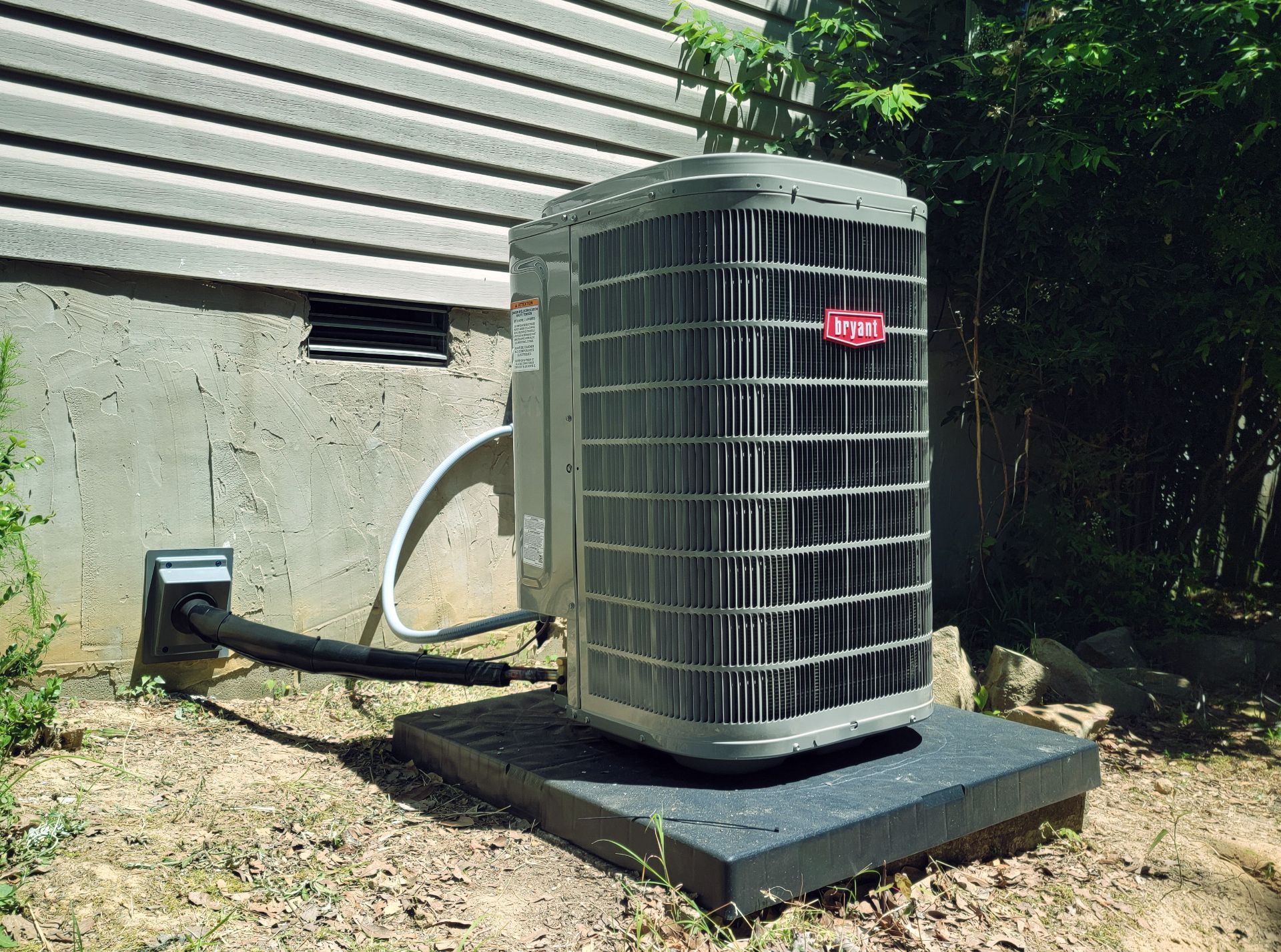 Outdoor air conditioner unit beside a house, with hoses and wiring visible on a concrete pad.