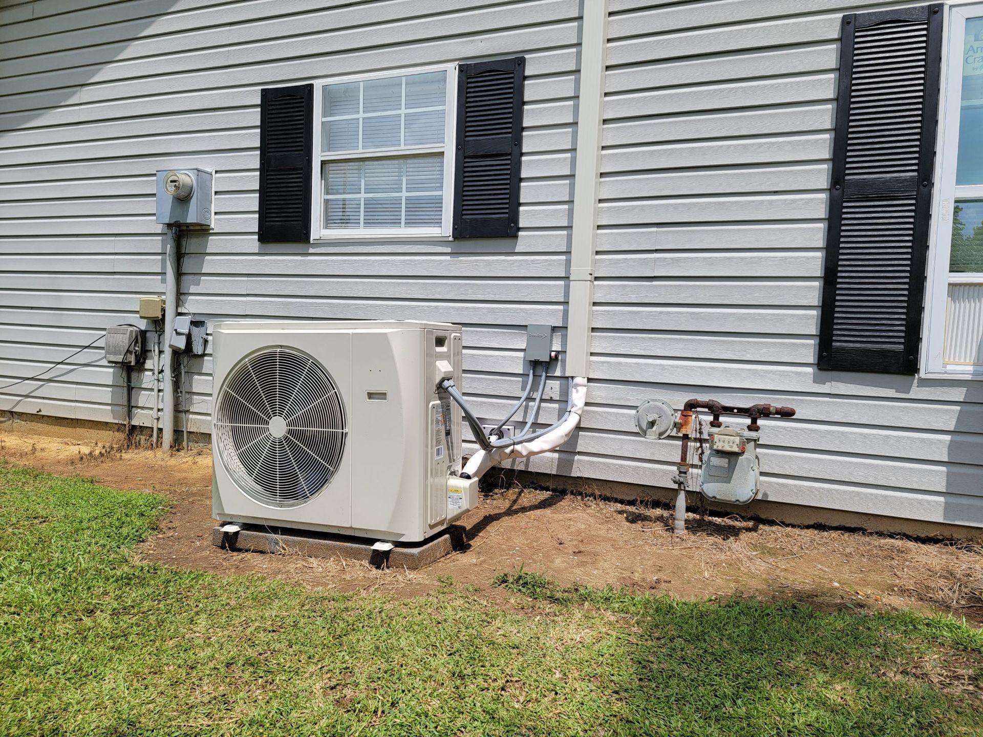 Outdoor AC unit beside a gray house with black shutters and exposed utility pipes.