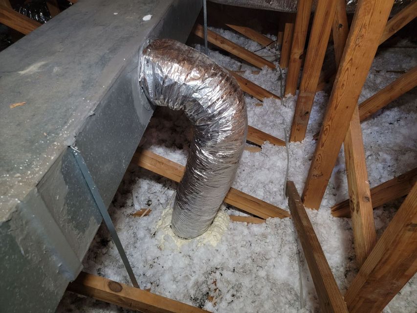 Flexible silver duct in a dusty attic beside wooden rafters and a sloped roof.