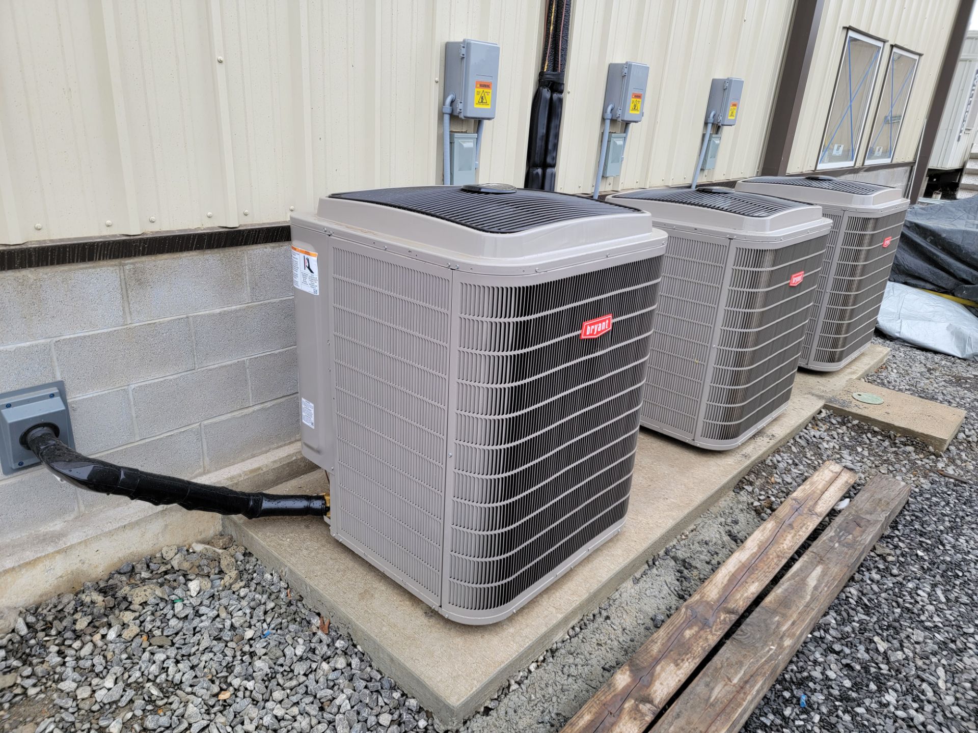 Three gray outdoor HVAC units on concrete pads beside a building wall, with gravel and lumber nearby