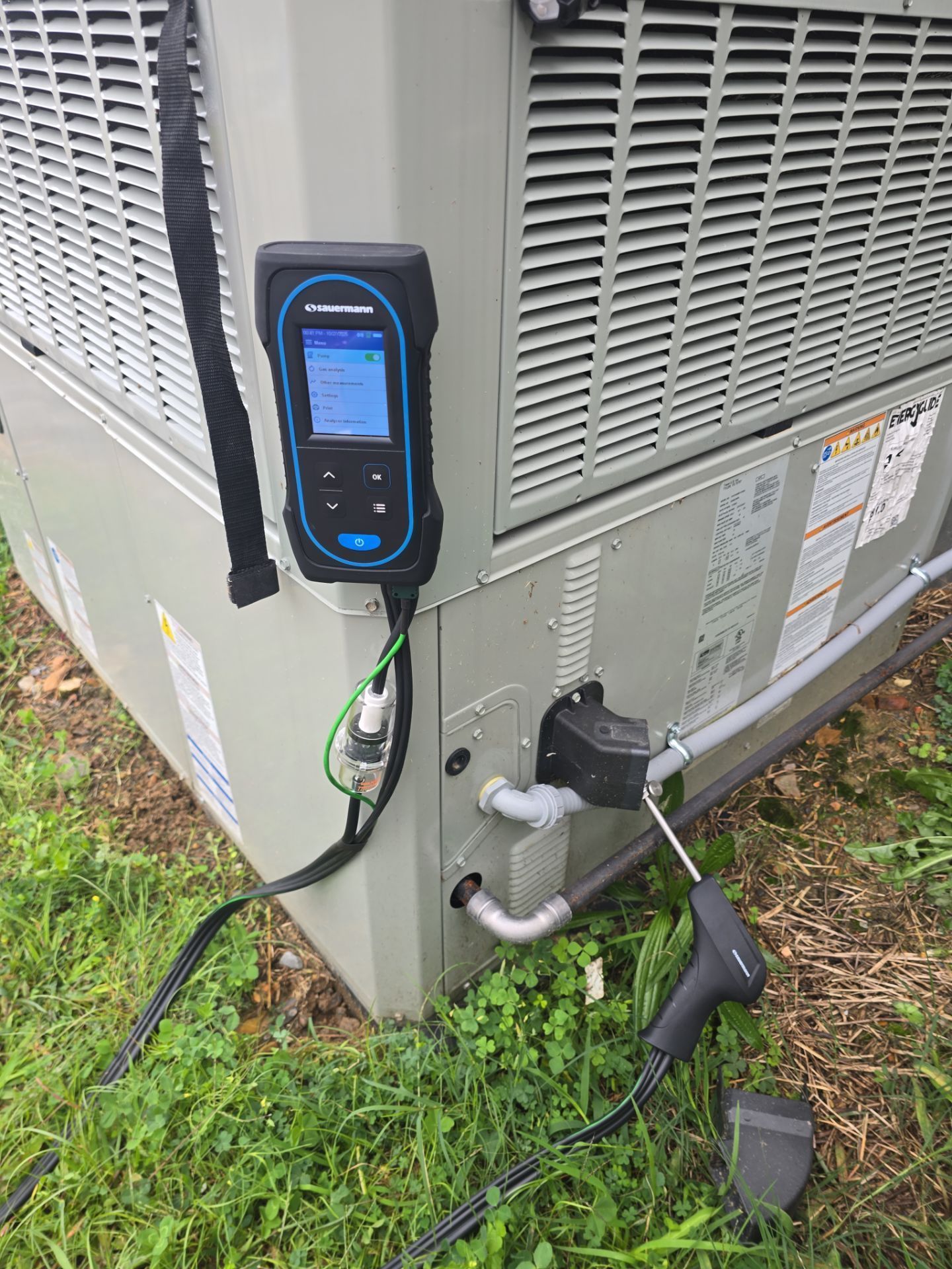 Portable diagnostic meter attached to an outdoor HVAC unit with hoses and cables on the ground