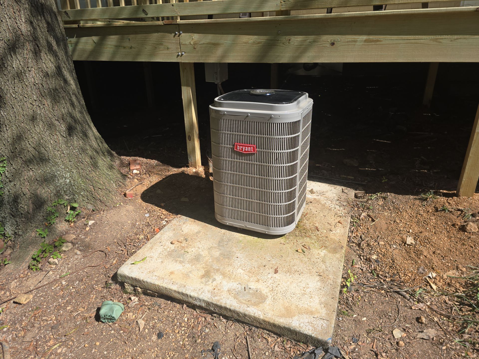 Outdoor air conditioner unit on a concrete pad beside a house deck and tree trunk.