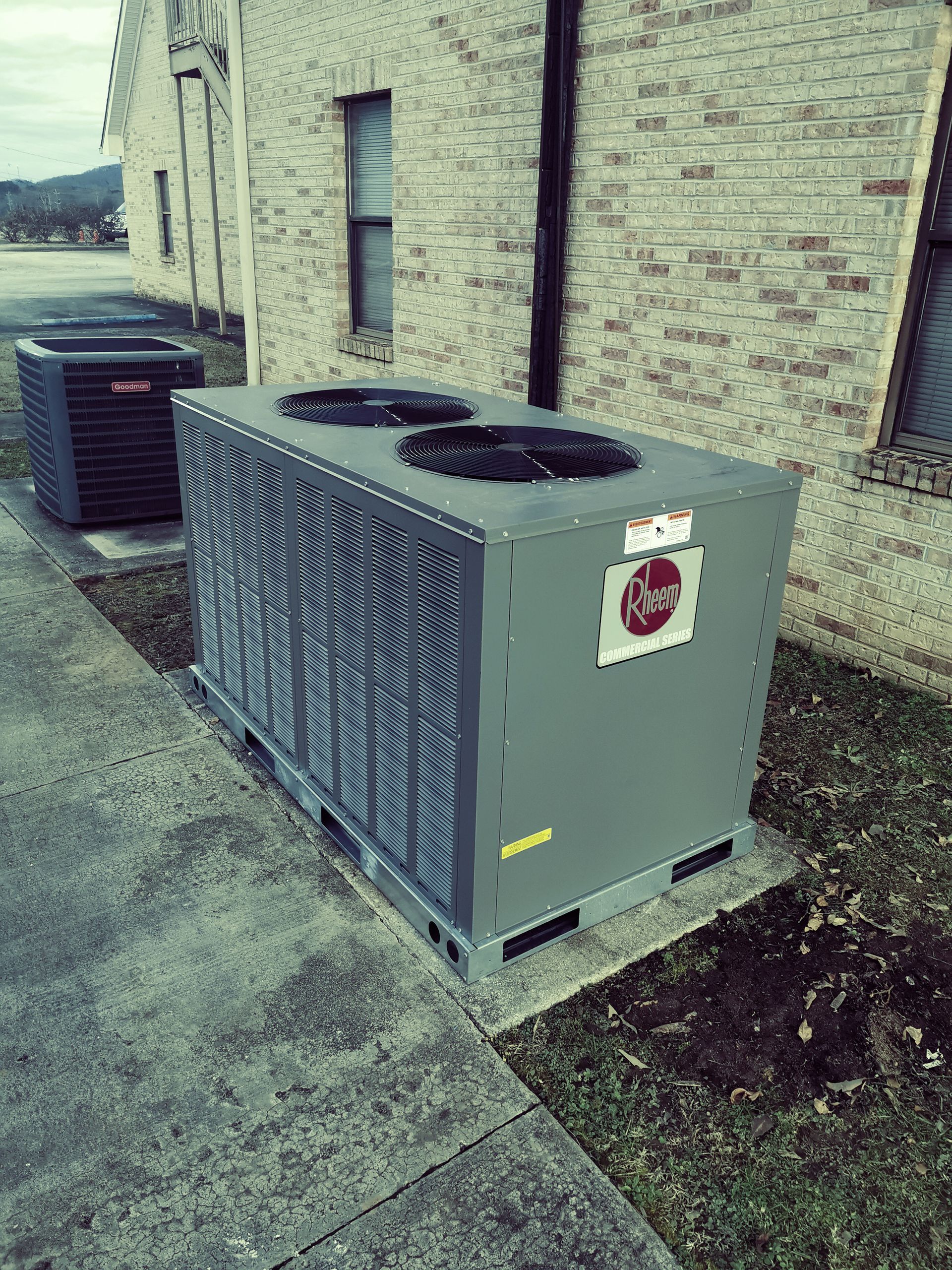 Gray HVAC unit beside a brick building on a sidewalk, with another unit in the background.