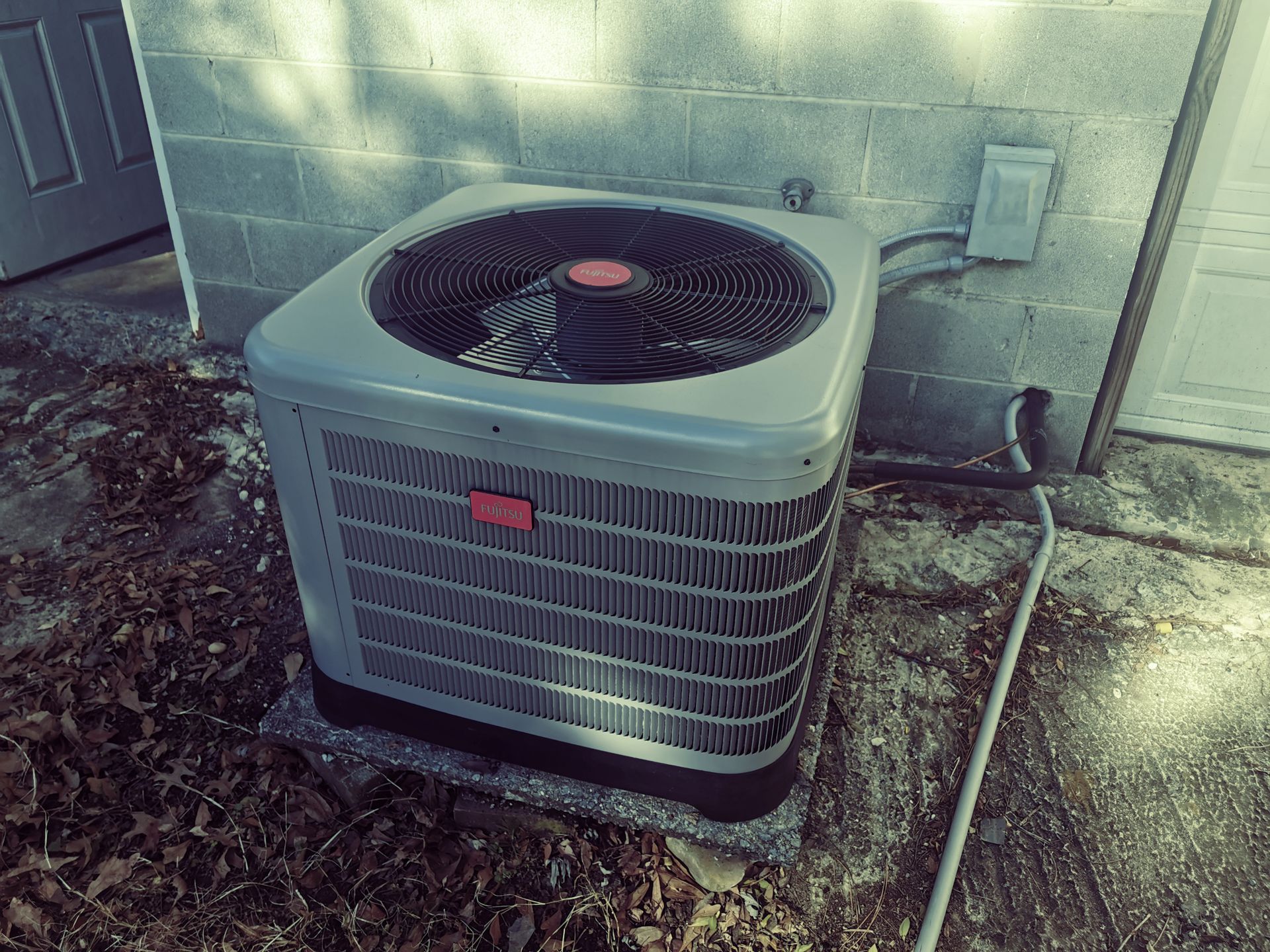 Outdoor air conditioner unit beside a house wall, with a fan and service hose attached