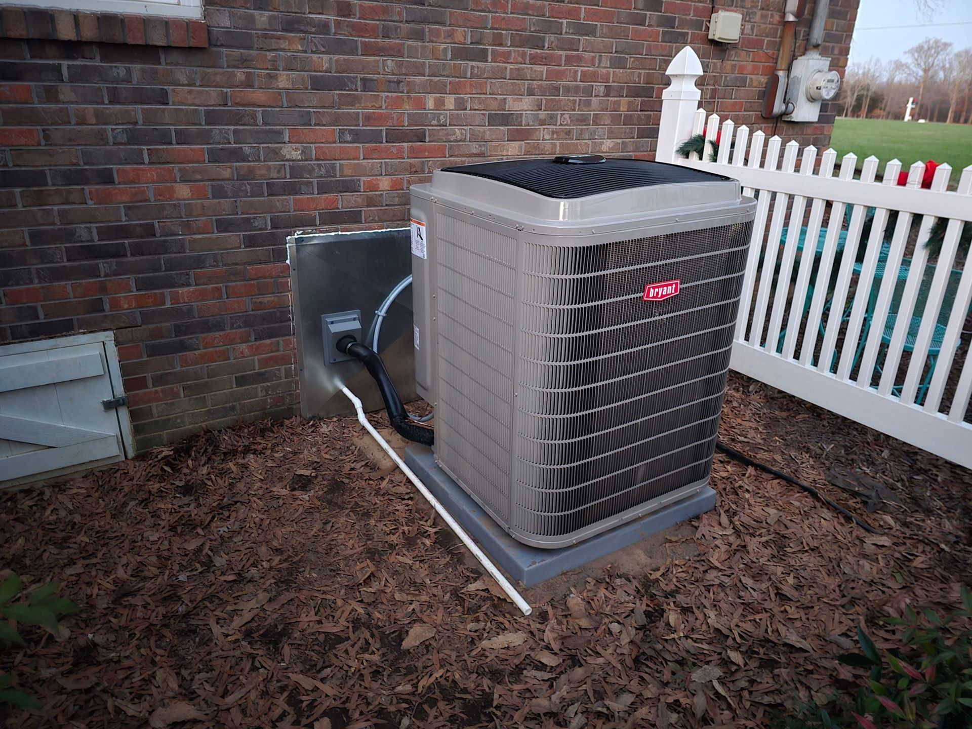 Outdoor air conditioner unit beside a brick wall and white fence, sitting on a mulch bed.