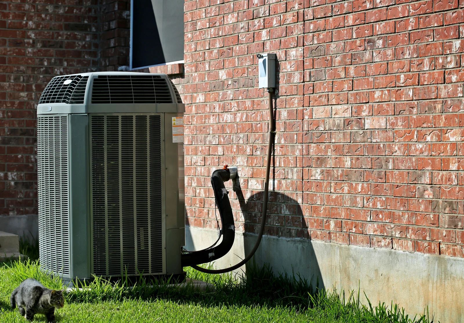 Outdoor air conditioner unit beside a red brick wall, with black hoses and green grass below.