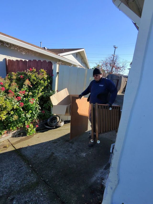 Person pushing a hand truck loaded with boxes along a narrow side yard beside a house