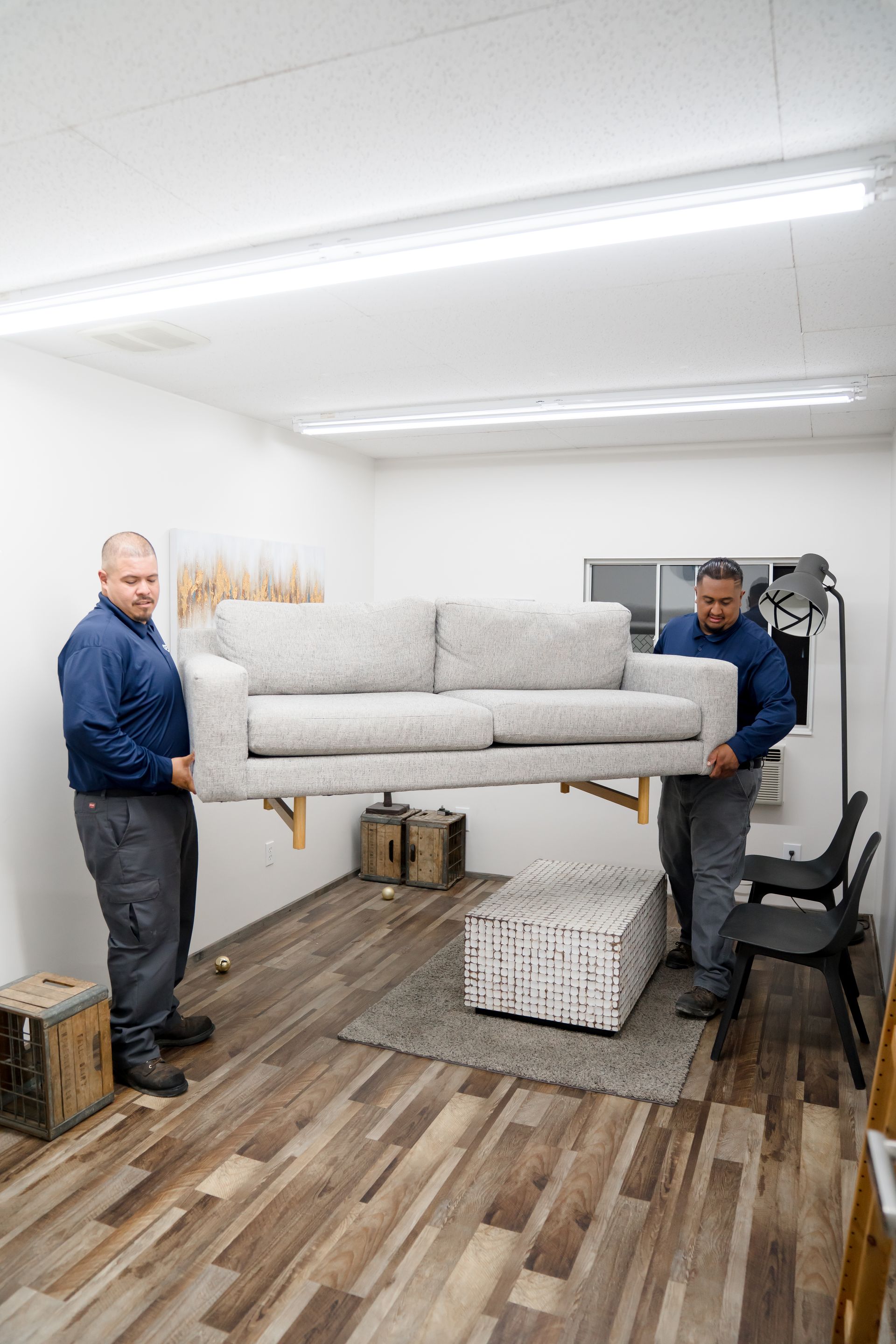 Two smiling men in matching navy shirts lift a gray sofa.