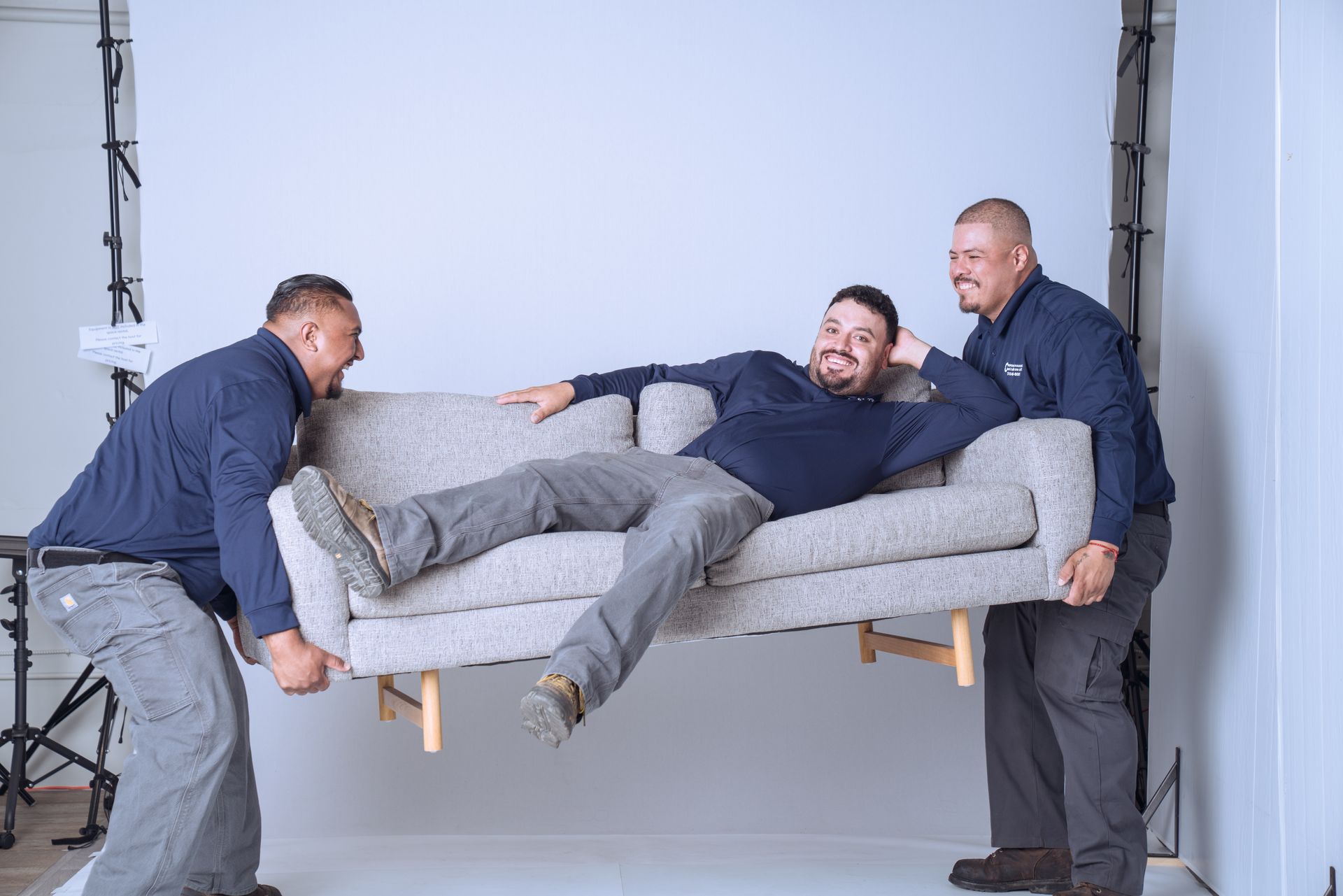 Two smiling men in matching navy shirts lift a gray sofa while a third man sits on the sofa.