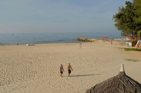 Two people walking on a sandy beach beside calm blue water and a rocky shoreline