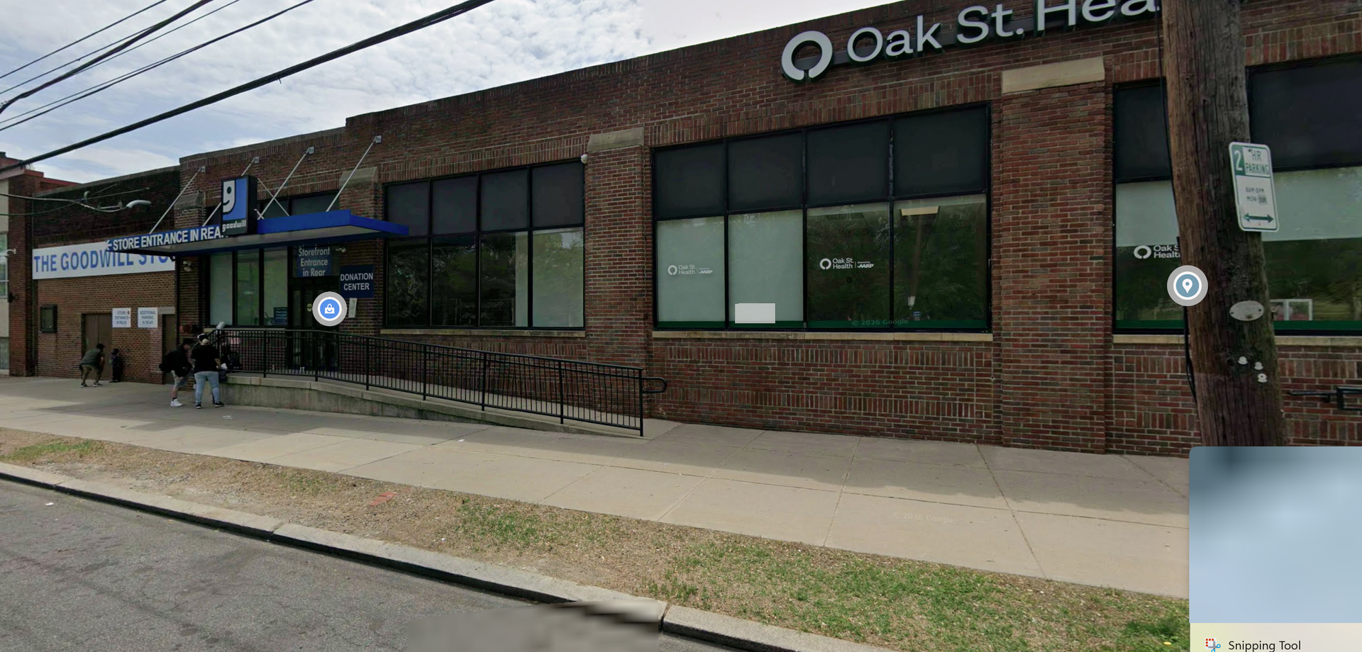 A street-level view of a brick Oak Street Health clinic building with a blue entrance awning and a wheelchair ramp.