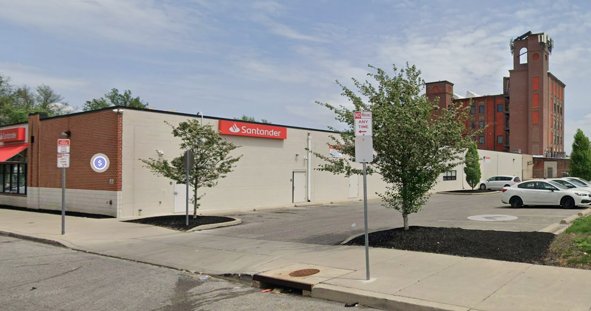A Santander Bank branch building with a parking lot, trees, and an industrial brick tower in the background.