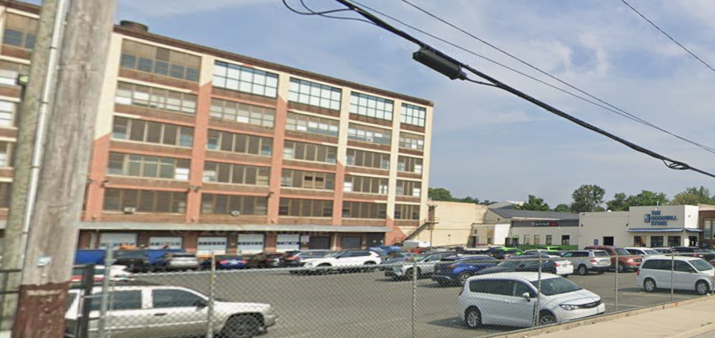 A large, multi-story brick building overlooks a paved parking lot filled with various cars on a sunny day.