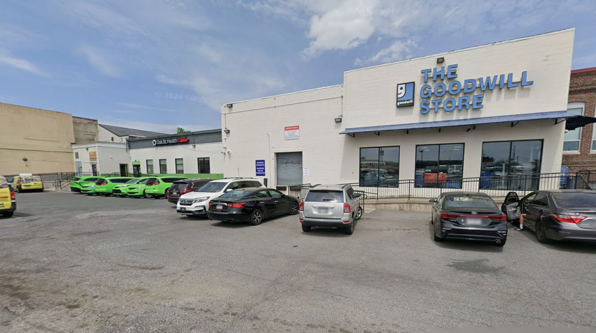 A Goodwill store building in a parking lot with several cars and green donation bins in front under a blue sky.