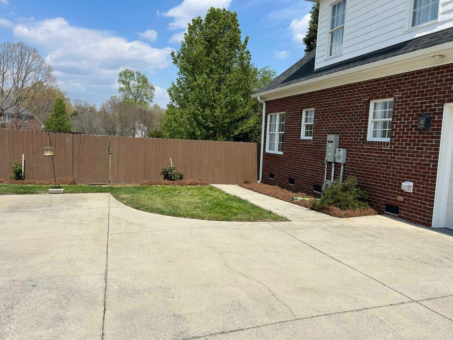 Concrete driveway beside a brick house, with a fenced backyard and a small grass patch under a blue sky.