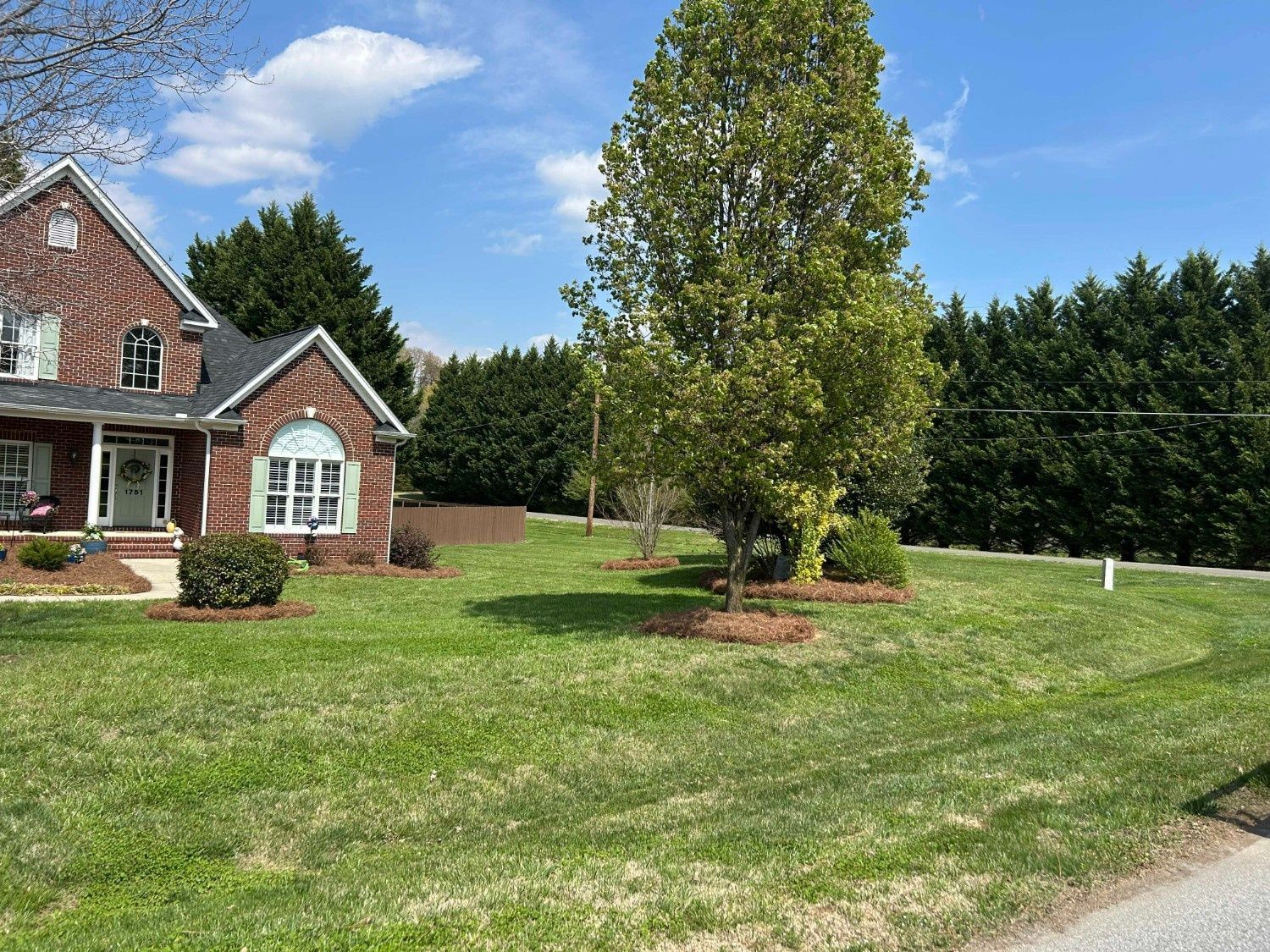 Brick house with white trim beside a large green lawn and a tall tree under a blue sky